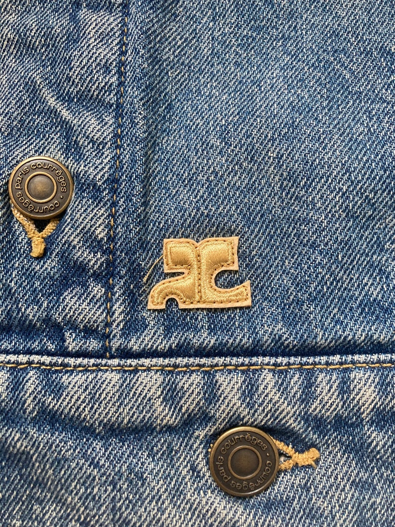 May include: A close-up of a blue denim jacket with two metal buttons that say "Paris Courr&egrave;ges" and a brown leather patch with the letter "C" in a stylized font.