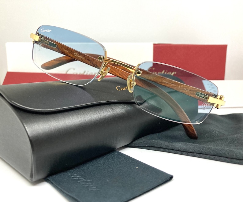 May include: A pair of Cartier sunglasses with a gold frame and light blue lenses. The frame is made of wood and has a Cartier logo on the side. The sunglasses come with a black case and a black cleaning cloth.