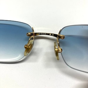 May include: Close-up of the gold-tone metal arms of a pair of blue gradient sunglasses. The arms are engraved with the numbers "5513 325" and "19".