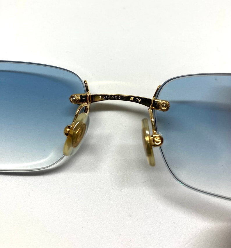 May include: Close-up of the gold-tone metal arms of a pair of blue gradient sunglasses. The arms are engraved with the numbers "5513 325" and "19".