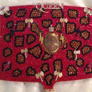 May include: A red, black, and gold hand mirror with a leopard print design. The mirror has a crown, lightning bolts, and axes on it. The word "QUEEN" is spelled out in rhinestones at the top of the mirror.
