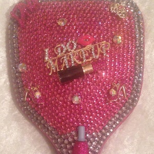 May include: A pink hand mirror covered in rhinestones with a gold "I Do Makeup" charm and a red lip charm.