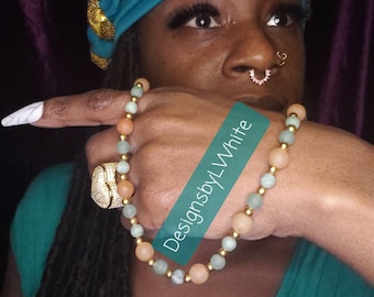 May include: A woman wearing a teal and gold turban with a beaded necklace and bracelet. The necklace and bracelet are made of light green, peach, and gold beads. The text "DesignsbyLWhite" is visible on the image.