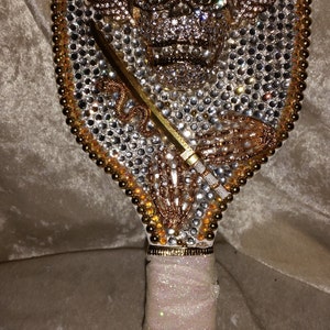 May include: A white hand mirror with a gold and silver skull design covered in rhinestones. The handle is wrapped in white fabric with gold trim.