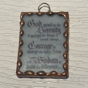 May include: A rectangular stained glass pendant with a copper-colored frame and a small loop at the top. The text reads "God grant me the Serenity... Courage... Wisdom..." in elegant script. The background is a light blue color.