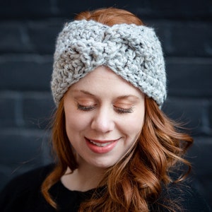 May include: A gray crocheted headband with a twisted knot design. The headband is worn by a person with long, red hair.