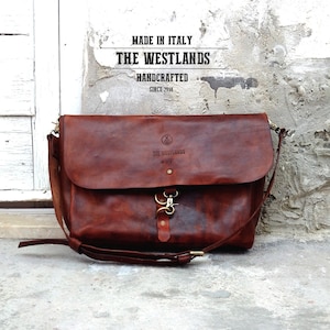 May include: A brown leather messenger bag with a flap closure and a buckle. The bag has a strap and a logo that says "The Westlands" and "Handcrafted Since 2014".