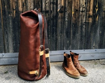 Könnte beinhalten: Eine braune Ledertasche mit schwarzem Riemen und Schnallen. Die Tasche liegt auf einer Holzoberfläche neben einem Paar braunen Lederstiefeln.