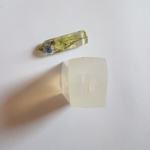 May include: A clear resin pendant with dried flowers and a square resin mold. The pendant is rectangular with blue and green flowers encased inside. The mold is a square shape with a small design inside.