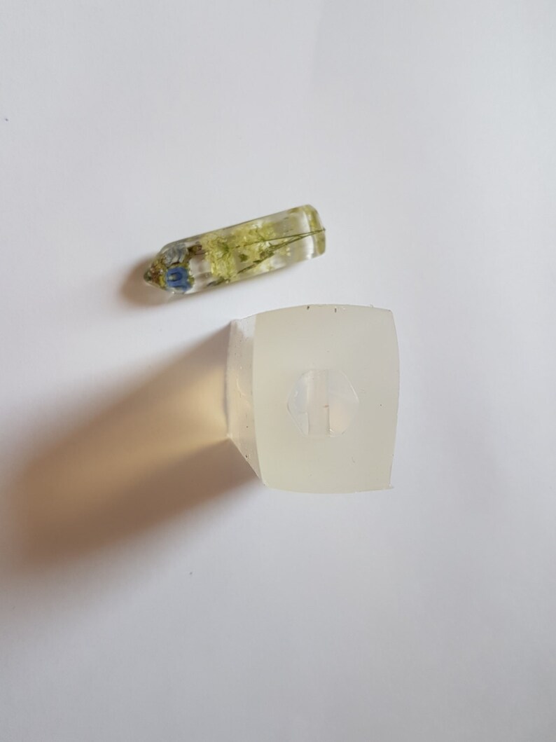 May include: A clear resin pendant with dried flowers and a square resin mold. The pendant is rectangular with blue and green flowers encased inside. The mold is a square shape with a small design inside.