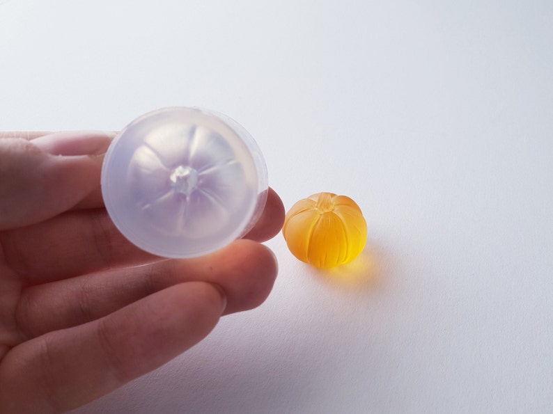 May include: A clear silicone mold shaped like a pumpkin, next to a small orange pumpkin-shaped resin piece.