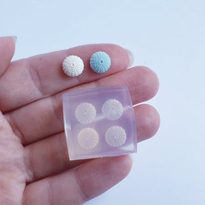 May include: A close-up of a hand holding a clear mold with four small, round, textured objects. Two finished objects, one white and one light blue, sit above the mold. The objects resemble miniature sea urchins, perfect for crafting.
