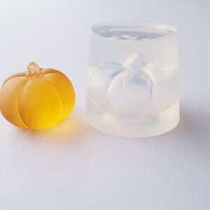 May include: A clear silicone mold shaped like a pumpkin, next to a small, amber-colored glass pumpkin.