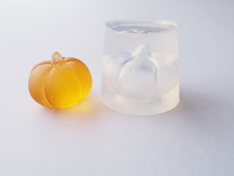 May include: A clear silicone mold shaped like a pumpkin, next to a small, amber-colored glass pumpkin.
