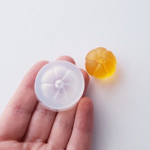 May include: A clear silicone mold shaped like a pumpkin with a small hole in the center. Next to the mold is a small, orange-colored pumpkin made of resin.