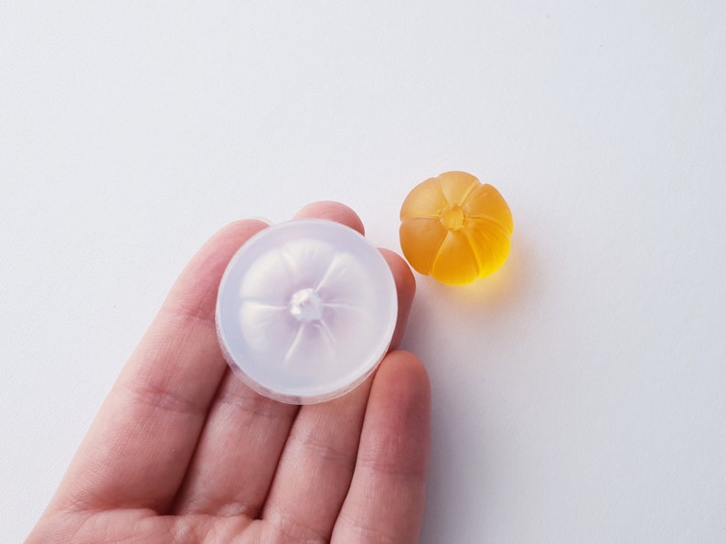 May include: A clear silicone mold shaped like a pumpkin with a small hole in the center. Next to the mold is a small, orange-colored pumpkin made of resin.