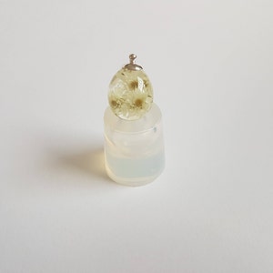 May include: A clear glass pendant with a silver cap and dried white flowers inside. The pendant is sitting on a white silicone mold.