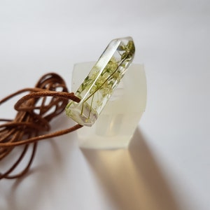 May include: A clear resin pendant with dried green and yellow flowers encased inside. The pendant is rectangular with a pointed end and is attached to a brown leather cord. The pendant is resting on a white surface.
