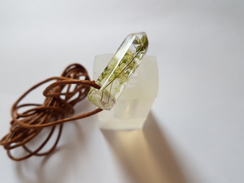 May include: A clear resin pendant with dried green and yellow flowers encased inside. The pendant is rectangular with a pointed end and is attached to a brown leather cord. The pendant is resting on a white surface.