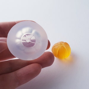 May include: A clear silicone mold shaped like a small orange, next to a small orange-colored resin casting.