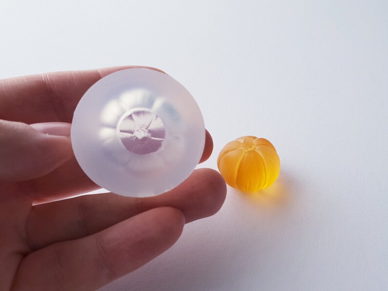 May include: A clear silicone mold shaped like a small orange, next to a small orange-colored resin casting.