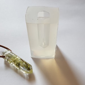 May include: A clear resin pendant with dried greenery inside, suspended on a brown leather cord. Next to it is a clear, rectangular silicone mold with a handle. The pendant is approximately 1.5 inches long.