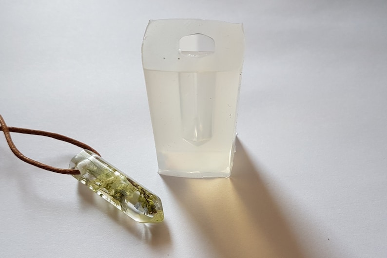 May include: A clear resin pendant with dried greenery inside, suspended on a brown leather cord. Next to it is a clear, rectangular silicone mold with a handle. The pendant is approximately 1.5 inches long.