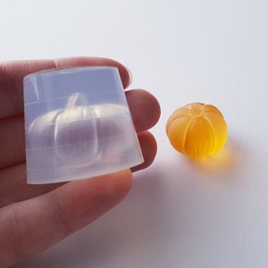 May include: A clear silicone mold shaped like a pumpkin, next to a small orange pumpkin made of resin.