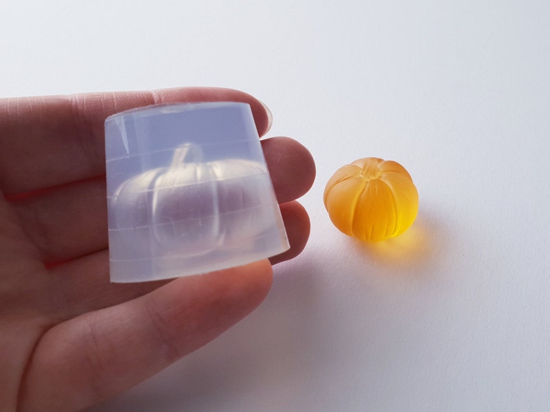 May include: A clear silicone mold shaped like a pumpkin, next to a small orange pumpkin made of resin.
