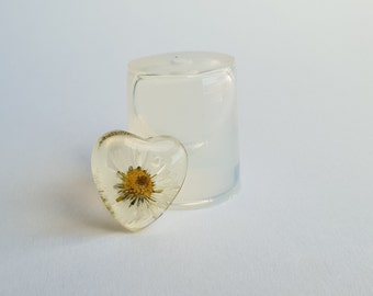 May include: A clear heart-shaped resin mold with a pressed flower inside. The flower has white petals and a yellow center.