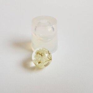 May include: A clear resin sphere with a dried white flower inside. The sphere is sitting next to a clear silicone mold.