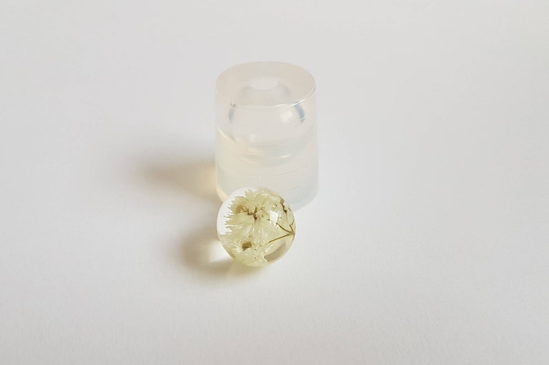 May include: A clear resin sphere with a dried white flower inside. The sphere is sitting next to a clear silicone mold.