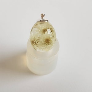 May include: A clear resin egg pendant with dried white flowers inside. The pendant has a silver bail for attaching to a chain or cord.