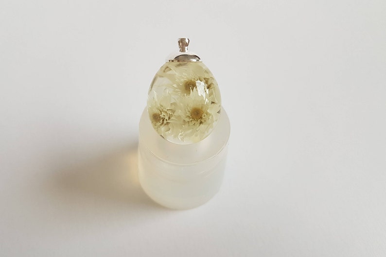 May include: A clear resin egg pendant with dried white flowers inside. The pendant has a silver bail for attaching to a chain or cord.