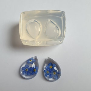 May include: Two clear resin teardrop-shaped pendants with blue forget-me-not flowers pressed inside. A silicone mold for making resin pendants is also shown.
