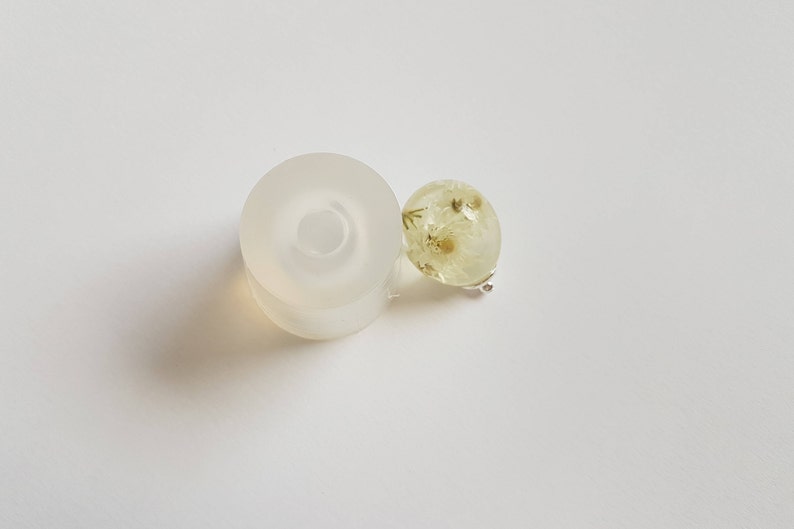 May include: A clear silicone mold for making resin jewelry, with a round, flat base and a raised ring in the center. Next to the mold is a small, round, clear resin pendant with dried white flowers inside.
