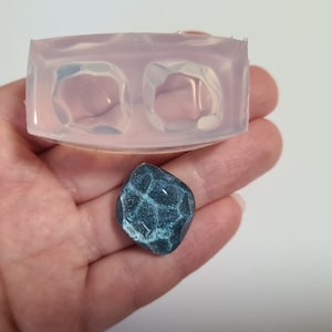 May include: A clear silicone mould with two cavities shaped like irregular gemstones. A teal blue resin piece with a textured surface is shown next to the mould.