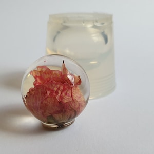 May include: A clear resin sphere with a dried pink flower inside. The sphere is sitting on a white surface next to a clear silicone mold.