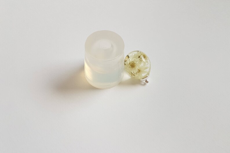 May include: A clear silicone mold for making round resin ornaments, next to a finished round resin ornament with dried white flowers inside.