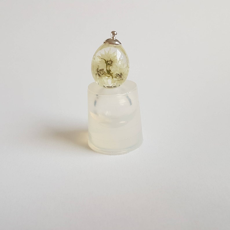 May include: A clear glass egg-shaped pendant with a silver cap and dried white flowers inside. The pendant is sitting on a clear silicone mold.