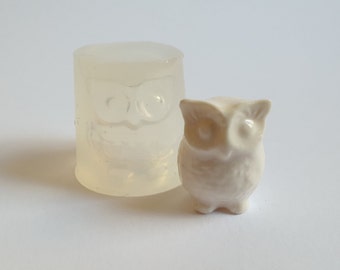 May include: A small, off-white ceramic owl figurine stands next to a clear, cylindrical silicone mold in the shape of an owl. The figurine has large eyes and a rounded body. The mold is translucent.