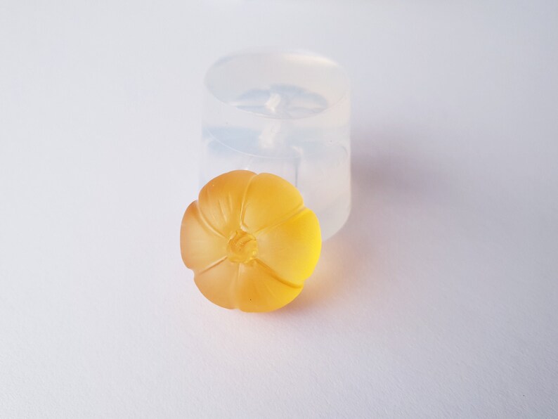 May include: A yellow resin flower charm with a detailed design. The charm is sitting next to a clear silicone mold.