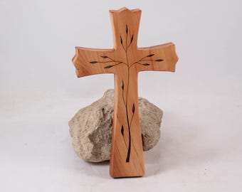 Könnte beinhalten: Ein Holzkreuz mit natürlichem Finish, mit einem geschnitzten Astdesign. Das Kreuz wird auf einem hellen Stein vor einem weißen Hintergrund präsentiert. Das Kreuz hat eine einzigartige Form mit einer leicht gebogenen Spitze.