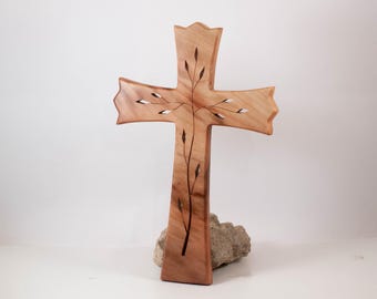 Könnte beinhalten: Ein hellbraunes Holzkreuz mit einem dunklen, eingelegten Zweigdesign, das sich vertikal von der Basis durch die Mitte erstreckt, mit kleinen blattförmigen Akzenten, die in die horizontalen Arme der Kreuzstruktur hineinragen.