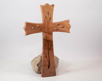 Könnte beinhalten: Ein hellbraunes Holzkreuz mit einem dunklen, eingelegten Zweigdesign und kleinen Blättern, die sich über die Mitte erstrecken. Das Kreuz hat ausgestellte Enden und wird an der Basis von einem rauen, hellgrauen Naturstein gestützt.