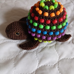 May include: A handcrafted crochet turtle with a brown body and shell. The shell is decorated with rows of colourful beads in shades of purple, blue, green, yellow, orange, and red. The turtle has small, dark eyes and is on a white quilted surface.