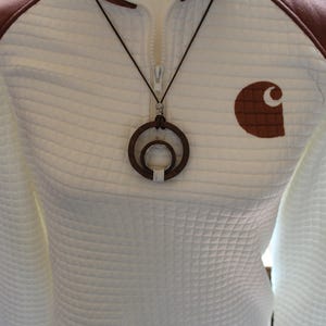 May include: A white quilted quarter-zip pullover with brown shoulder patches and a brown Carhartt logo on the chest. A dark brown leather cord necklace features a circular pendant with a smaller silver ring nested inside the dark wood frame.
