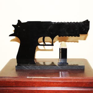 May include: A black silhouette of a handgun and a bullet, displayed on a wooden base. The gun is detailed with a textured slide and trigger. The bullet is gold and black. The base is a dark, polished wood.