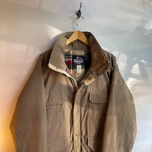 May include: A tan-colored jacket with a high collar and multiple front pockets. The jacket has a zipper and button closure. The interior lining features a plaid pattern. The jacket is hanging on a wooden hanger.