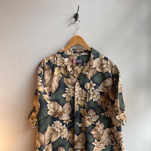 May include: A short-sleeved shirt with a floral design. The shirt has a dark green base with large beige and white flowers and leaves. It is suspended from a wooden hanger.